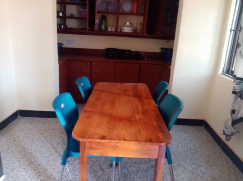 A wooden dining table is placed at the center, surrounded by four teal chairs. In the background, cabinetry is visible, displaying a collection of kitchenware and utensils on open shelves. A well-lit environment is created by the natural light filtering through the nearby window.