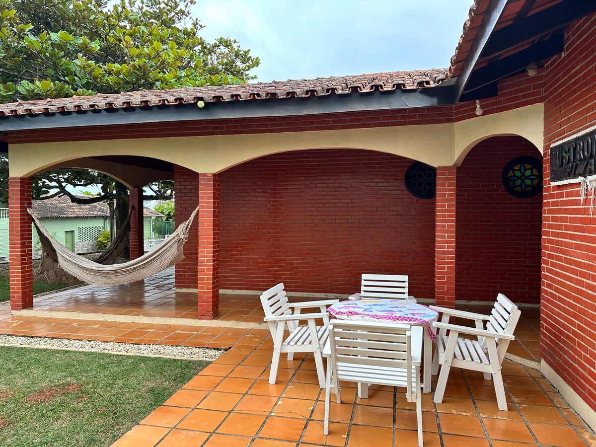 An outdoor area features a spacious brick patio, with a round table surrounded by four white chairs. A hammock is suspended between two columns, and lush greenery is visible in the background. The roof is adorned with traditional tiles.