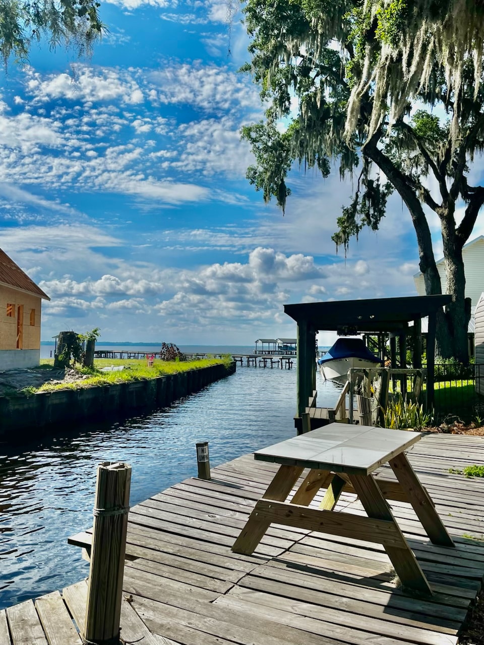 A serene view of the St. Johns River is captured from a wooden dock. A picnic table is positioned nearby, surrounded by lush greenery. The sky features scattered clouds, reflecting tranquility, while a glimpse of waterfront structures is visible in the background.