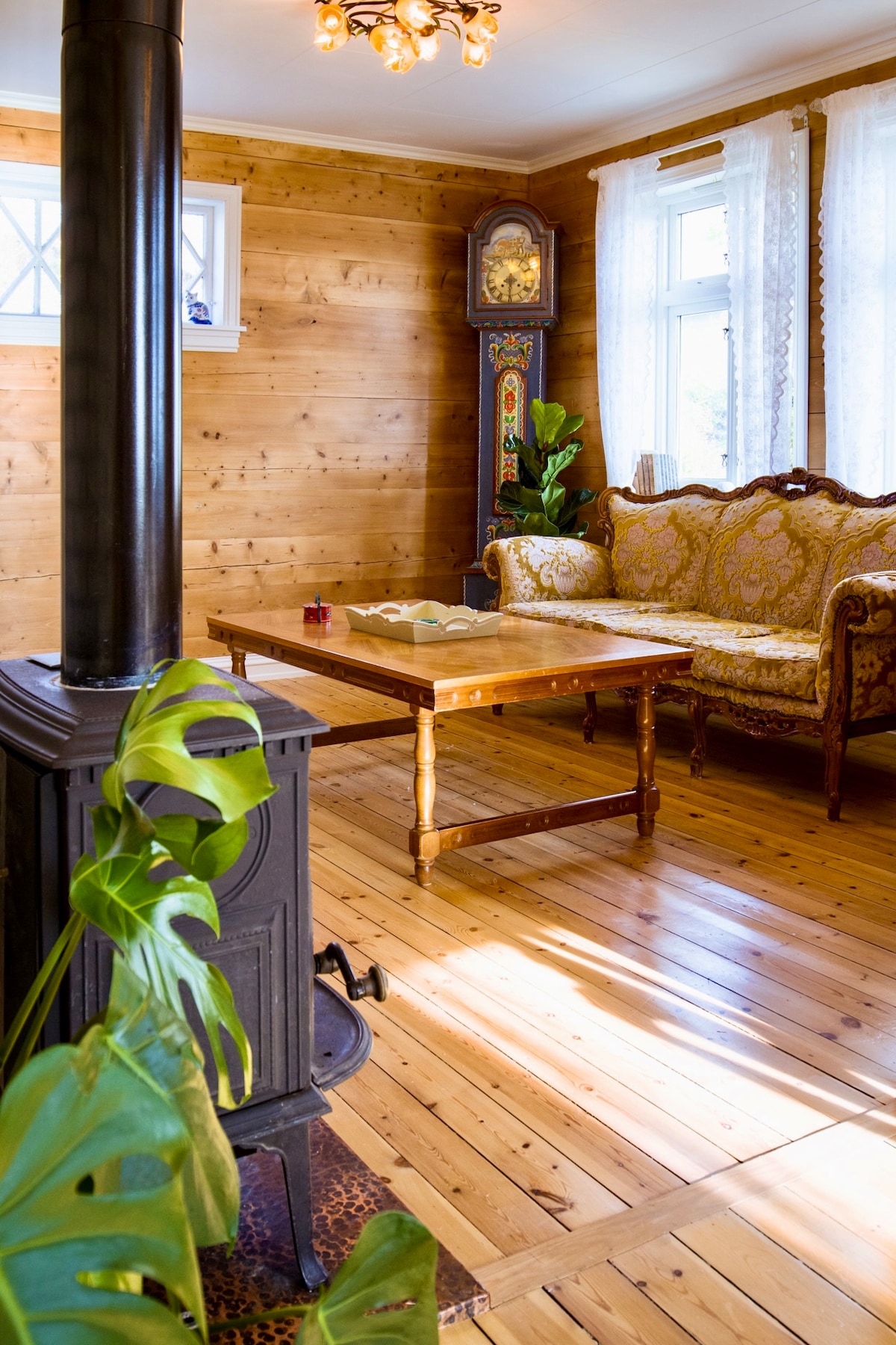 A charming sitting area features warm wooden walls and a polished wooden floor. An ornate sofa sits across from a wooden table, with a green plant in the foreground. Sunlight filters through sheer curtains, illuminating the space.