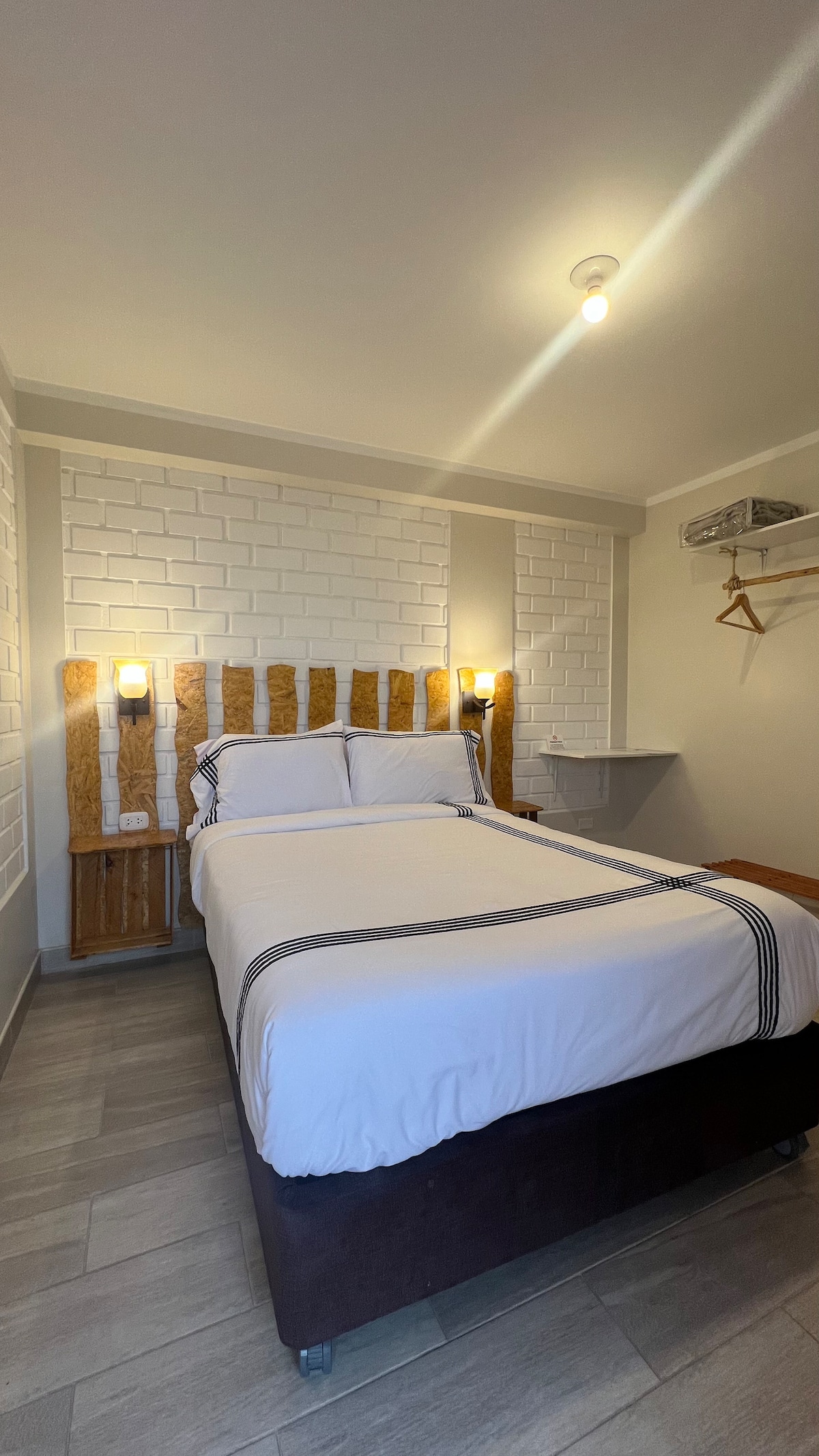 A cozy sleeping area is presented with a compact bed framed by wooden headboard details. Soft lighting is provided by wall sconces beside the bed. A small shelf and hanging space are visible, contributing to the room's functionality.