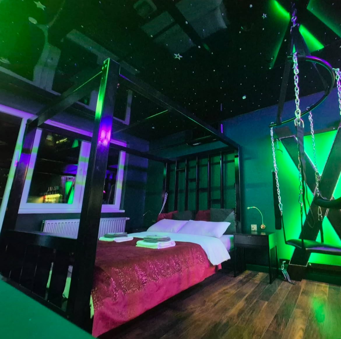 A four-poster bed is centrally positioned in a darkly painted room, complemented by green lighting that creates an ethereal atmosphere. The ceiling resembles a starry night sky, while a suspended swing adds a unique design element. Natural light is allowed in through a window.