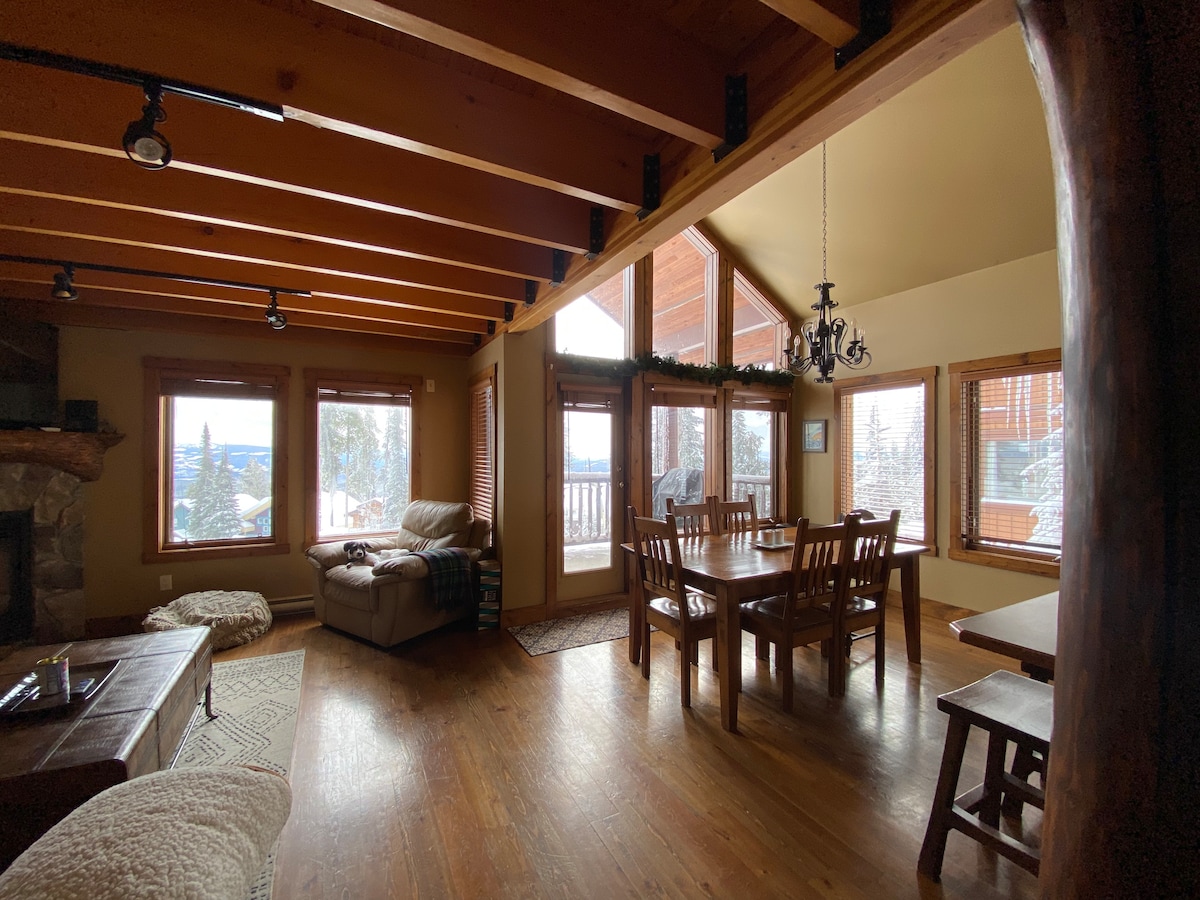 An open-concept living and dining area features warm wood tones and ample natural light from large windows. A rustic stone fireplace sits alongside a comfortable seating area, while a dining table accommodates multiple guests. Exposed wooden beams enhance the spacious atmosphere.