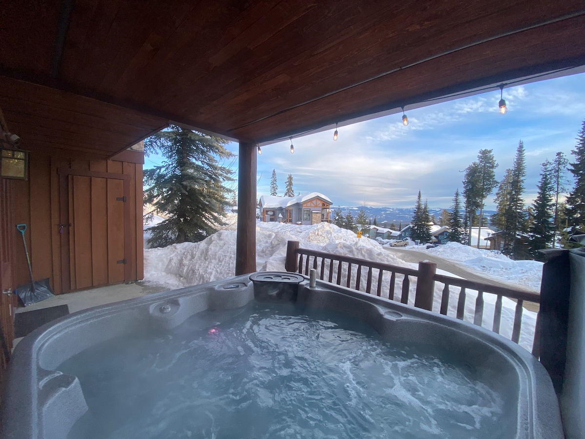 A hot tub is nestled under a covered deck, surrounded by wooden railings. The view features snow-covered slopes and evergreen trees under a cloudy sky, with gentle evening light reflecting off the water's surface.