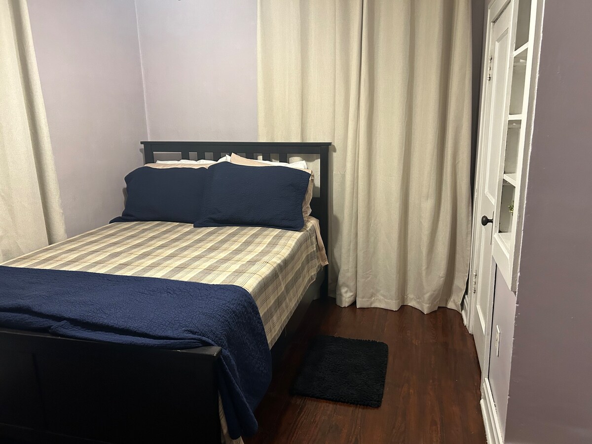A cozy bedroom features a black bed frame with a neatly made bed, adorned with two pillows and a striped blanket. Light-colored curtains frame a window, while a plush black rug lies on the hardwood floor, enhancing the room's comfort.
