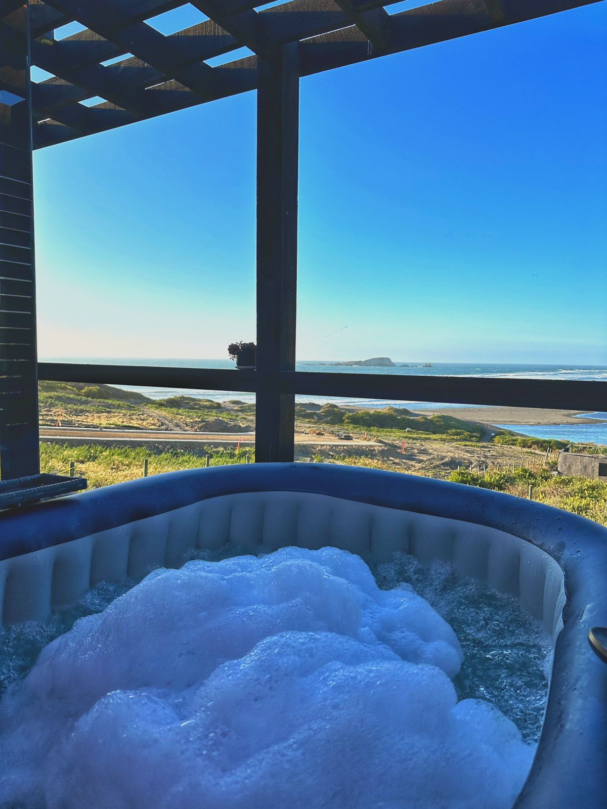 A personal electric hot tub is displayed, filled with bubbles, positioned on a terrace. Large windows frame a clear view of the ocean and coastal landscape, allowing natural light to fill the space.