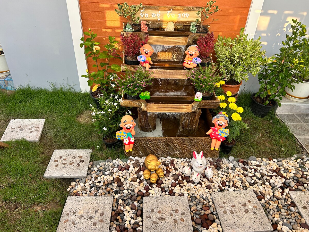 A serene outdoor area features a tiered water fountain surrounded by decorative pots and colorful plants. Small figurines are positioned alongside the fountain, enhancing the natural setting. Pebbled pathways meander through the greenery, creating an inviting space for relaxation.