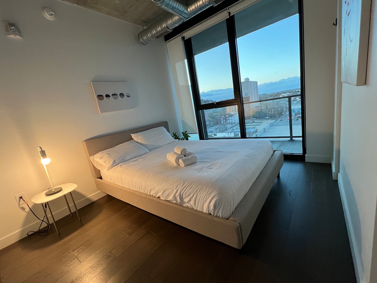 A cozy bedroom features a spacious bed with crisp white linens, accompanied by neatly arranged towels. Natural light filters through the floor-to-ceiling windows, revealing a view of the city and mountains beyond. A small side table with a lamp adds to the minimalist decor.