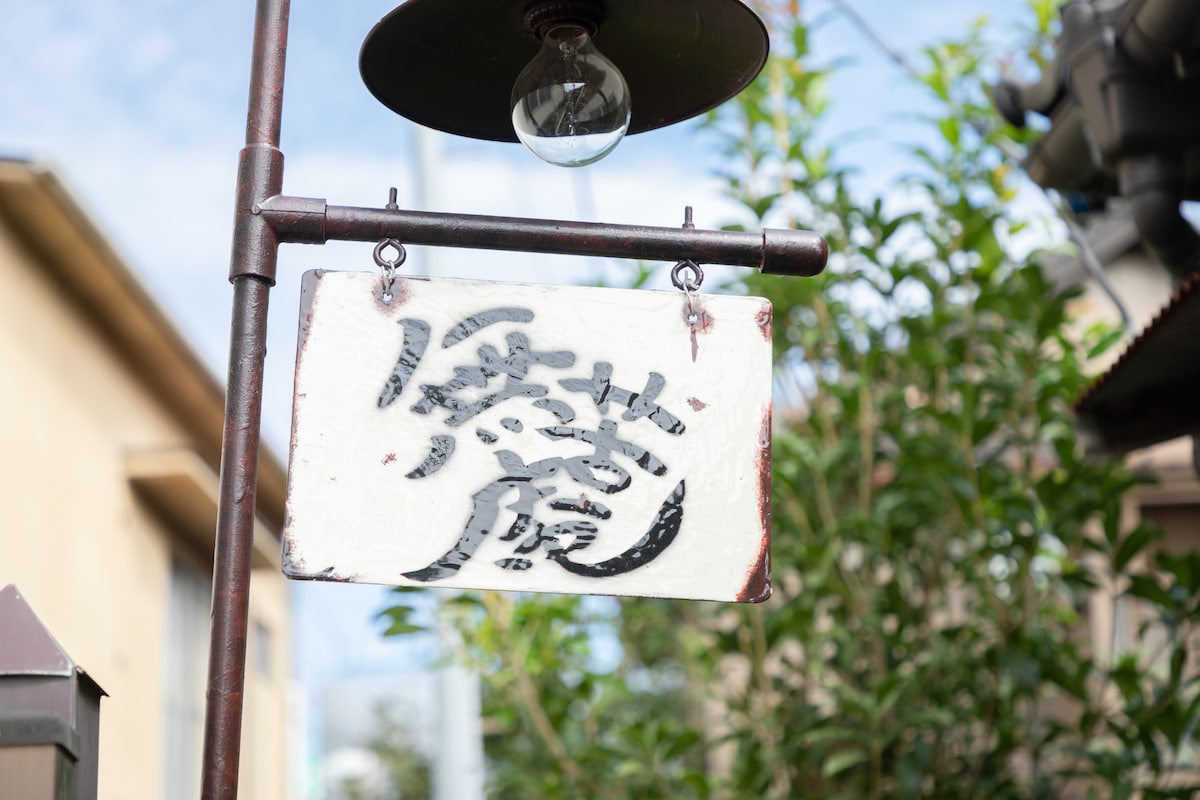 A weathered sign featuring traditional Japanese calligraphy hangs from a decorative metal arm. The backdrop includes lush green foliage and a clear blue sky, creating a serene and inviting entrance to the accommodation.