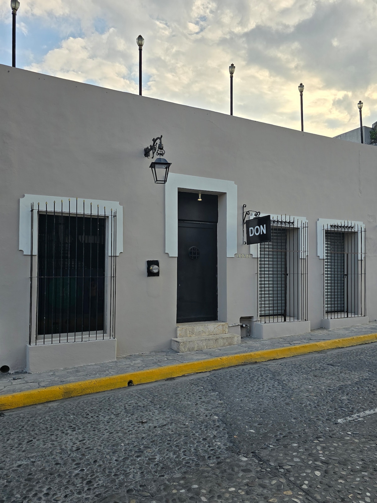 The exterior of the hotel showcases a charming colonial-style building. The smooth, light-colored walls are complemented by white window trims and a contrasting dark door. A prominent sign with the name 'DON' is visible, along with traditional wall-mounted lanterns, set against the backdrop of a cobblestone street.