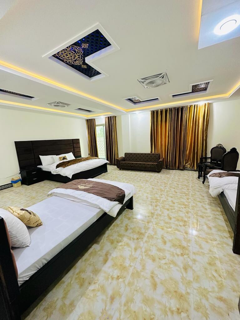 A spacious room features two sleek beds and a comfortable sofa, all arranged on a light-colored tiled floor. Elegant dark curtains frame a window, enhancing the natural lighting. Decorative ceiling elements add interest to the modern design, creating a calming atmosphere.