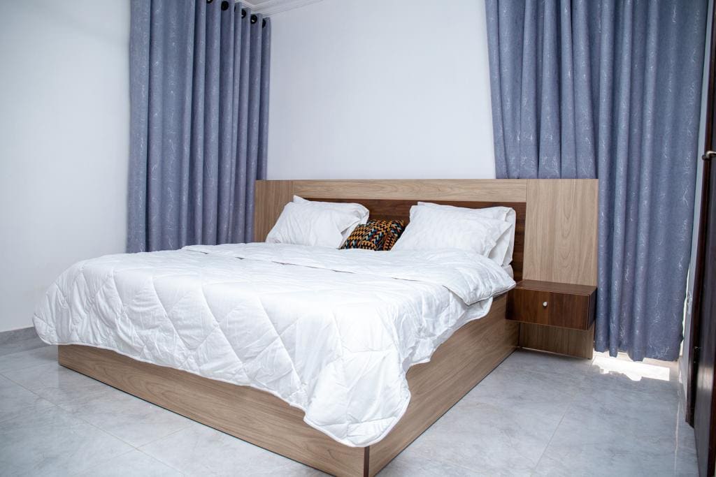 A spacious sleeping area is equipped with a large bed featuring a light quilt and decorative pillows. The wooden headboard complements the soft grey-blue curtains that frame the window, while tiled flooring enhances the room's overall brightness.