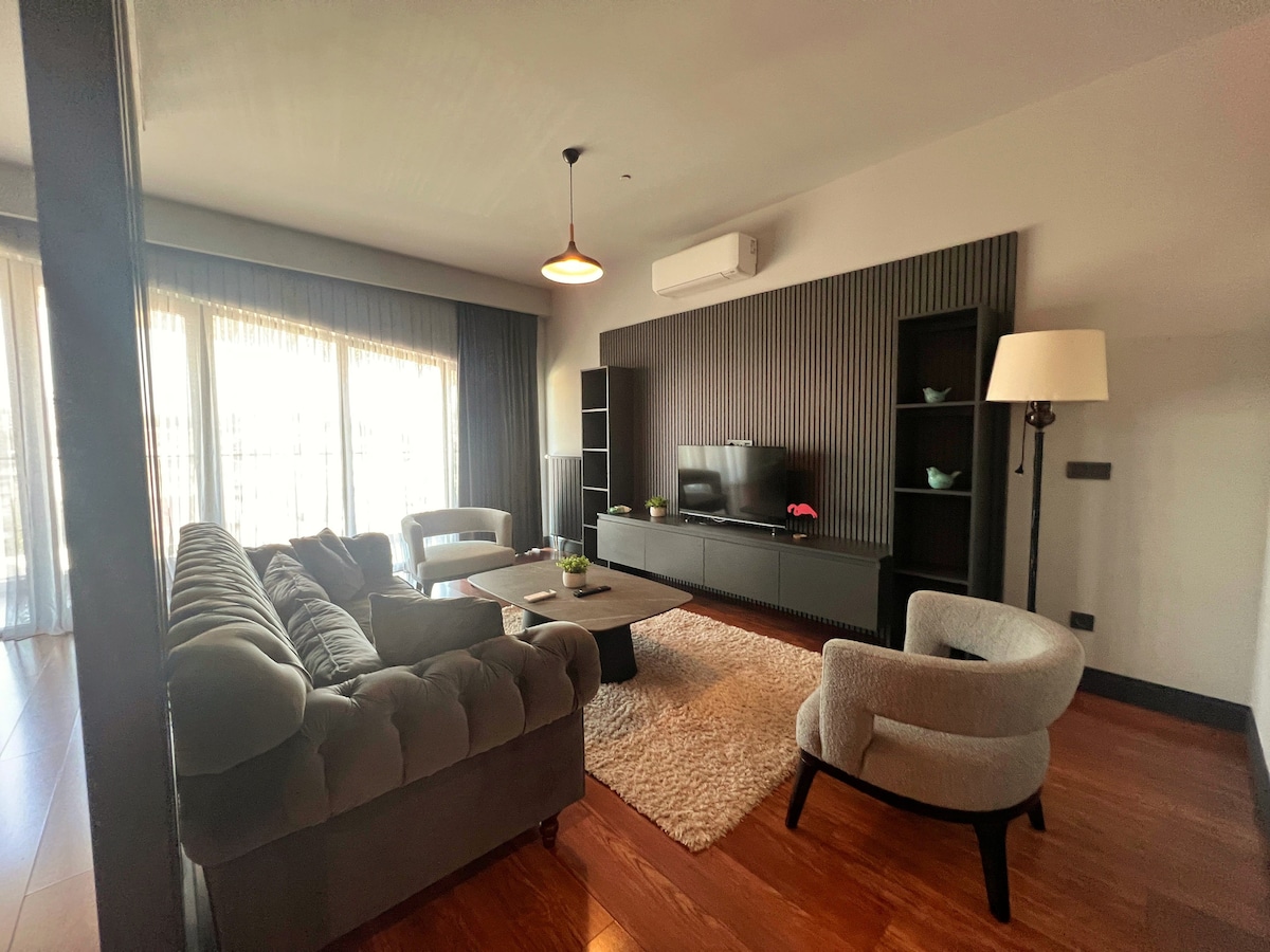 A spacious living area is furnished with a plush sectional sofa and modern armchairs. A sleek television unit against a textured wall holds a flat-screen TV. Natural light floods the room through expansive windows, highlighting the contemporary decor and a soft rug underfoot.