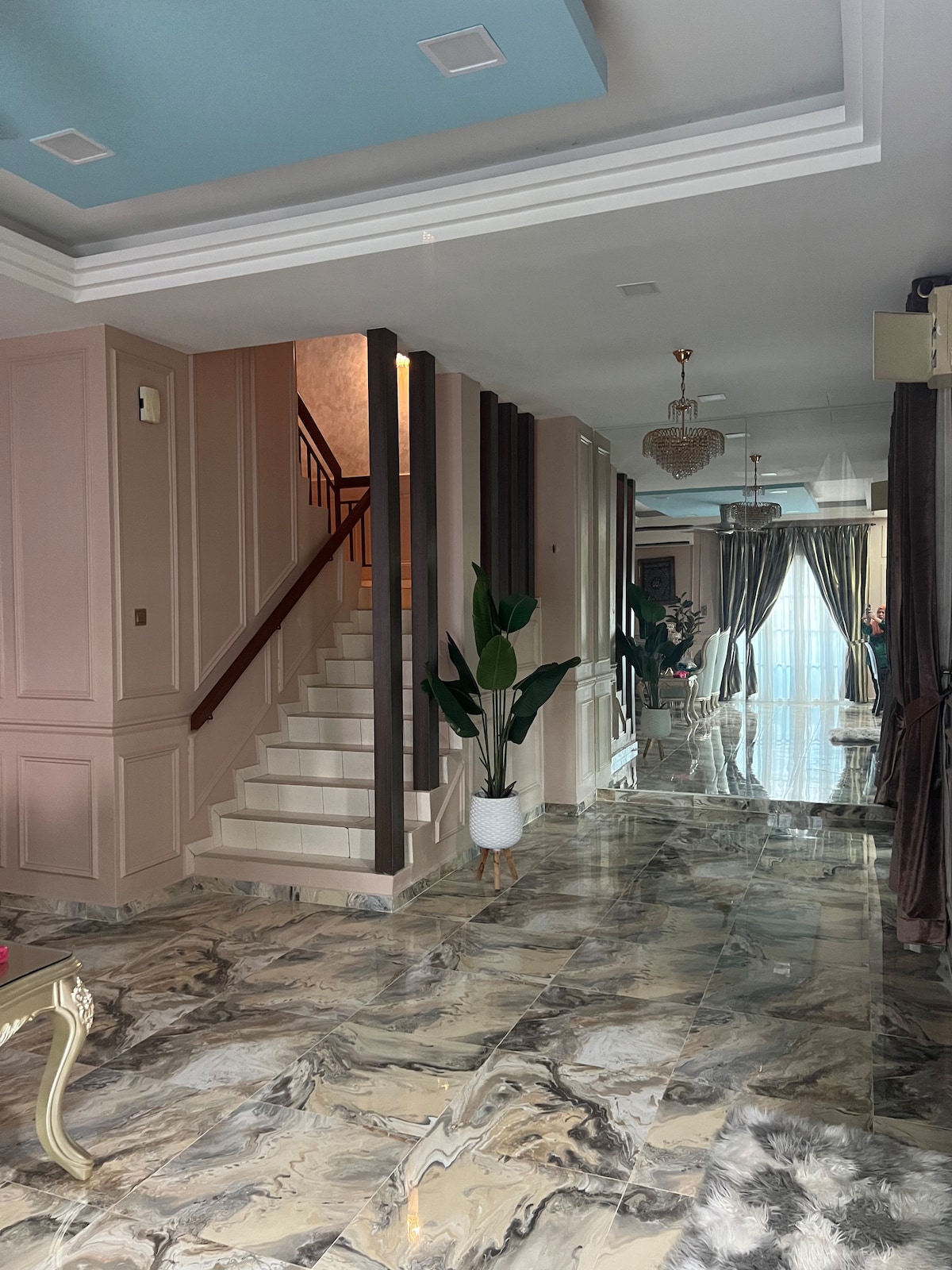 A spacious hallway presents decorative marble flooring that reflects natural light. The soft-colored walls are accented with subtle detailing, while a staircase leads to the upper level. Decorative plants are placed strategically, enhancing the welcoming atmosphere of the entryway.