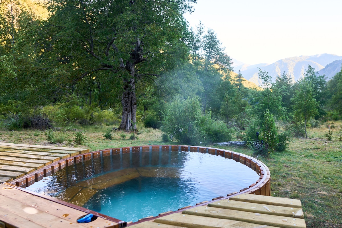 A wooden hot tub is set on a deck, surrounded by lush greenery and trees. Mist subtly rises from the water, enhancing the serene natural landscape filled with mountains in the background.