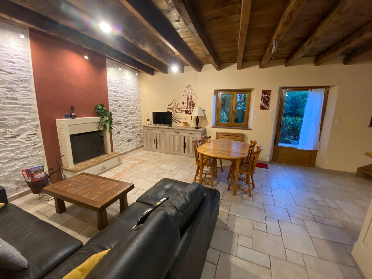 A cozy living area features a stone fireplace as a focal point, complemented by wooden beams above. A dark sofa faces a wooden entertainment unit, with a table and chairs arranged nearby. Natural light enters through a window, enhancing the space's inviting feel.