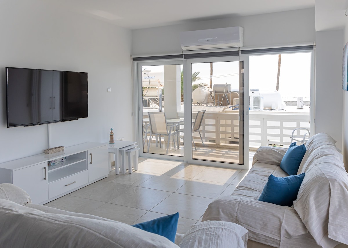 A bright living area features two white sofas adorned with blue cushions, complemented by a wall-mounted television. Large sliding glass doors provide access to a balcony with outdoor seating, enhancing the space's openness and connection to the sea views.