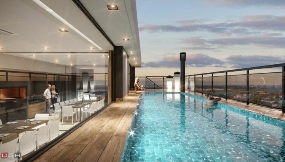 A rooftop area showcases a shimmering pool bordered by sleek glass railings. The wooden deck contrasts beautifully with the tranquil water. In the background, a dining space is visible through expansive windows, featuring white tables and chairs, highlighting an inviting ambiance.