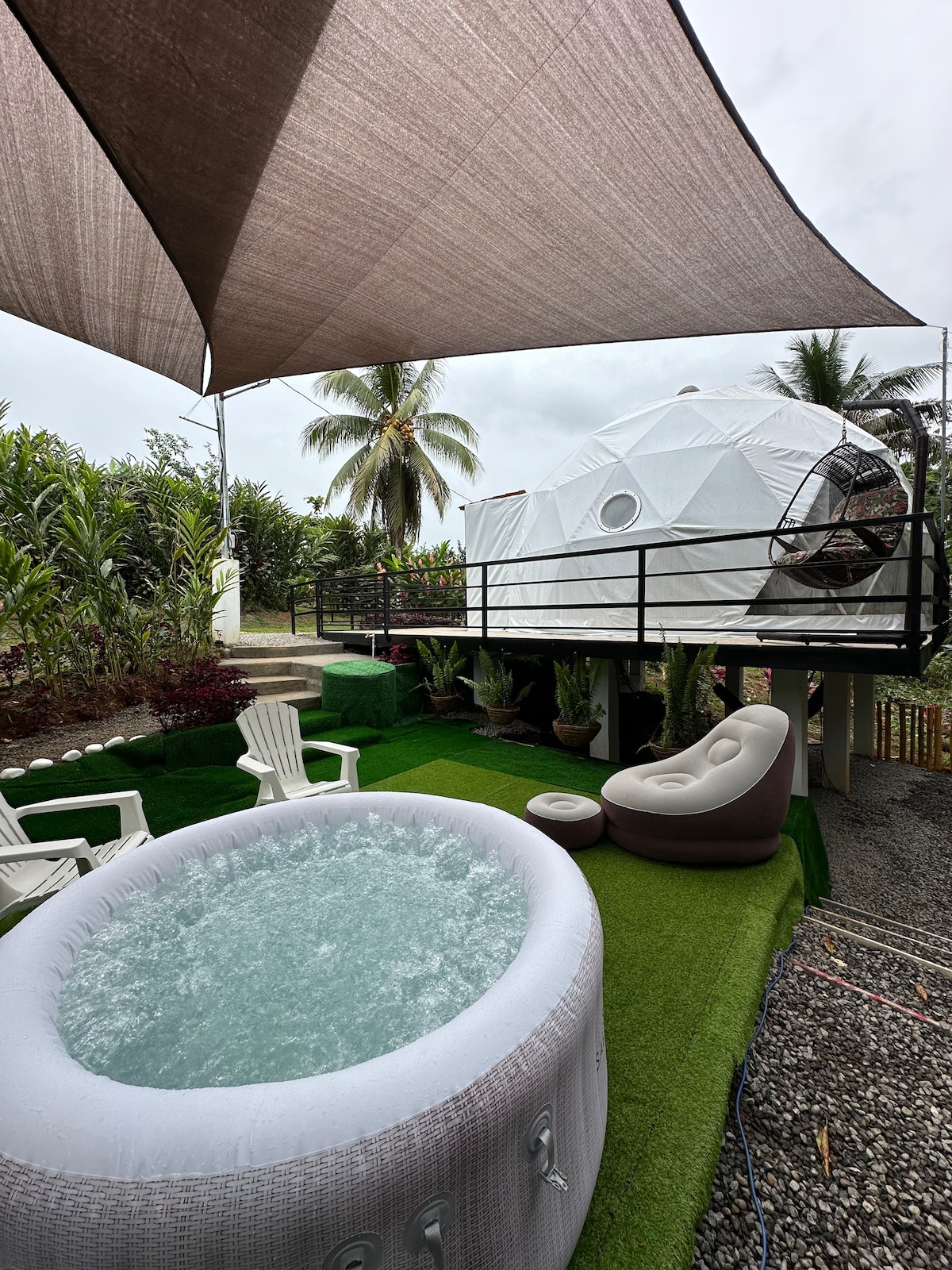 An outdoor area features a circular hot tub surrounded by comfortable seating arrangements. A shaded dome structure stands above, complemented by lush greenery and palm trees in the background. A layer of artificial grass adds a touch of nature and relaxation to the space.