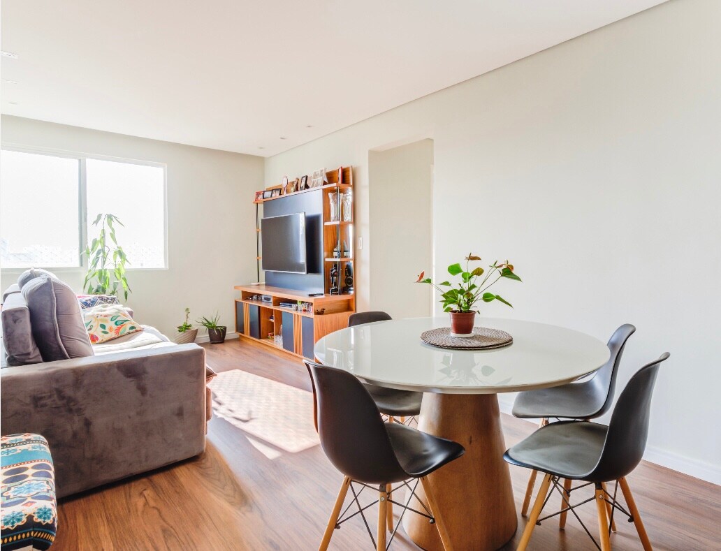 A bright living space features a spacious sofa adorned with colorful cushions. A modern round dining table with four black chairs sits on polished wood flooring. Natural light streams through large windows, illuminating a TV unit against the wall, complemented by indoor plants.