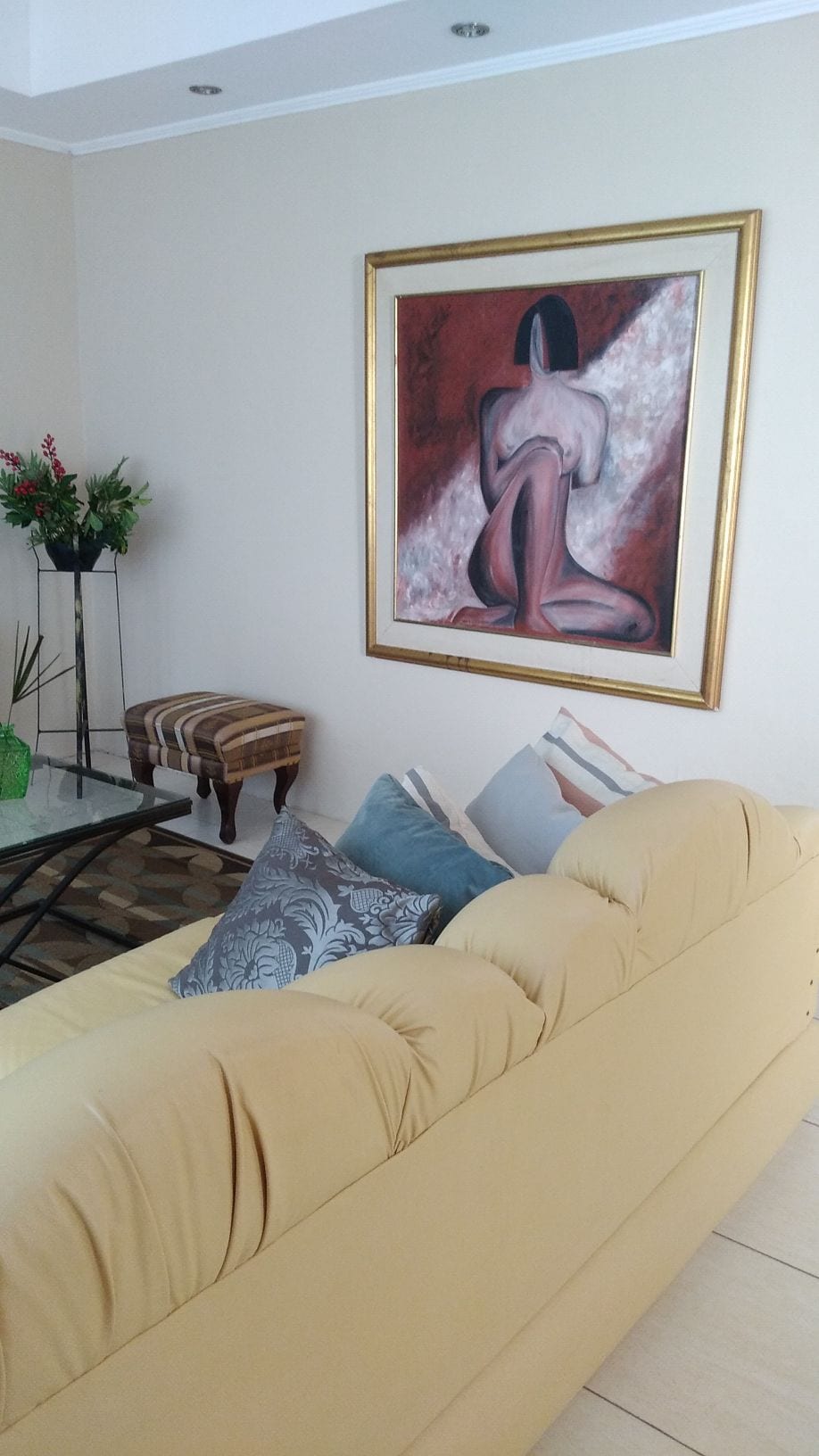 A light-colored sofa is positioned in front of a glass coffee table, with decorative pillows arranged on it. A framed painting of a figure is displayed on the wall, and a side table with a vase of flowers adds a touch of greenery to the space.