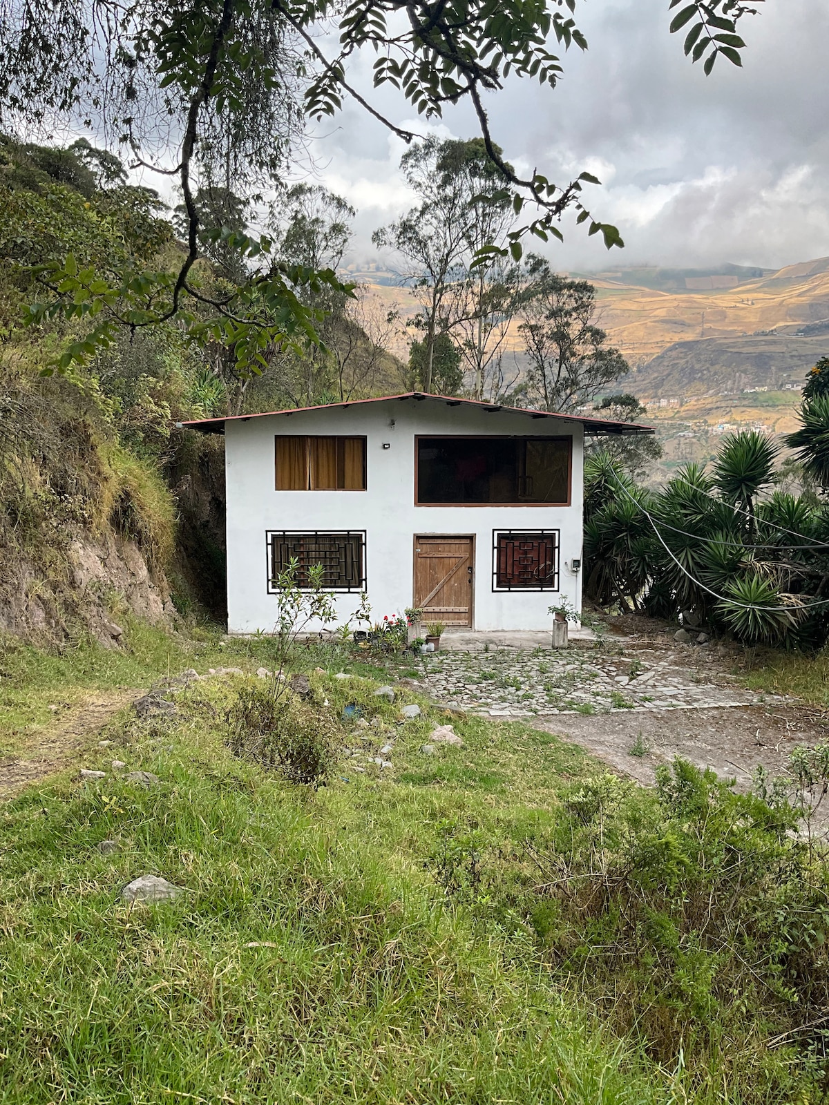 A two-story house with a white exterior is surrounded by greenery and rugged terrain. Large windows offer views of the landscape, while a stone pathway leads to the entrance. The natural surroundings create a peaceful, rural atmosphere.