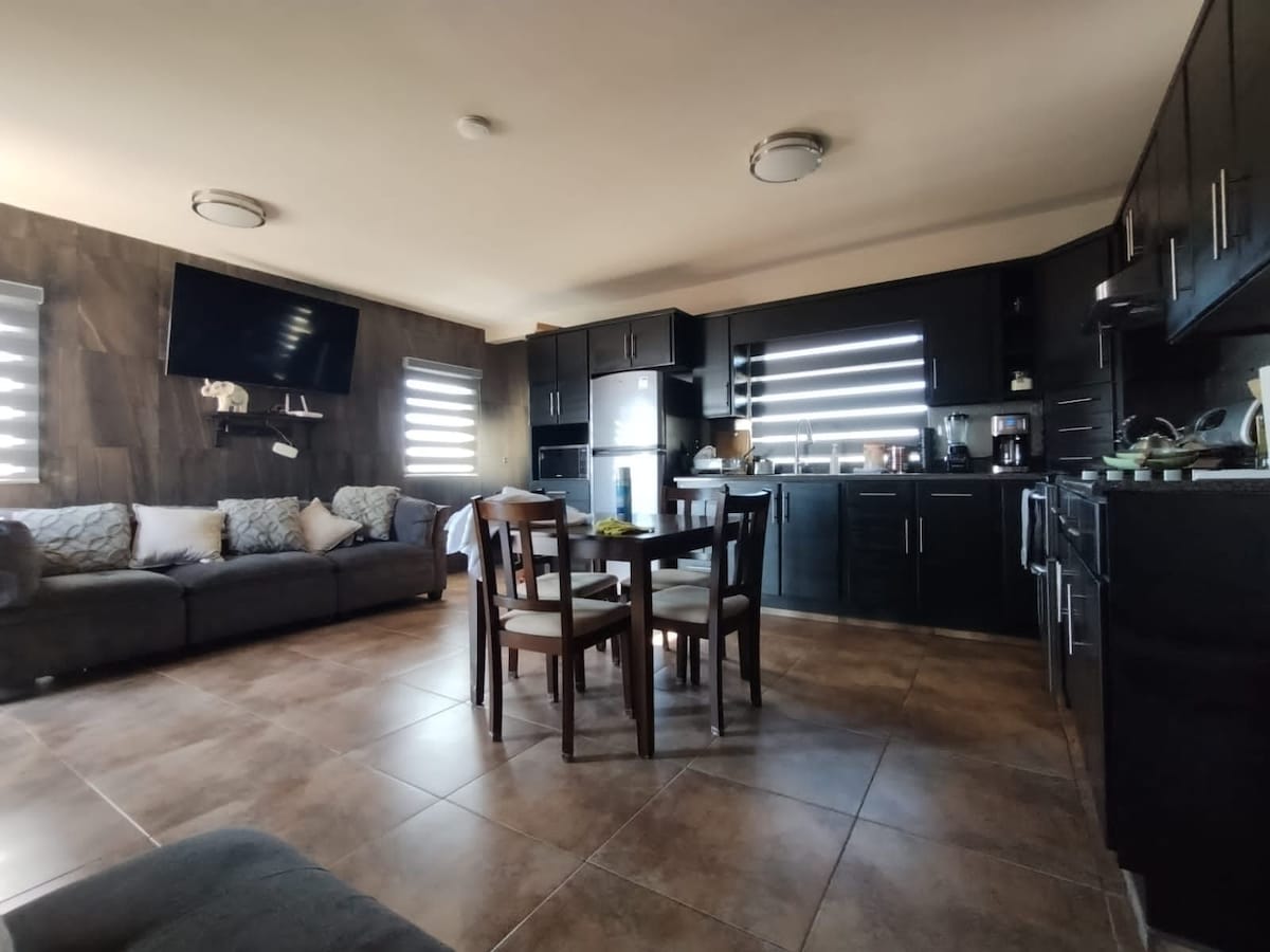 A modern kitchen and living area feature dark cabinetry and sleek appliances. A spacious sofa is positioned against the wall, complemented by decorative cushions. A dining table with four chairs occupies the center, while a large flat-screen television is mounted on the wall, contributing to functionality.