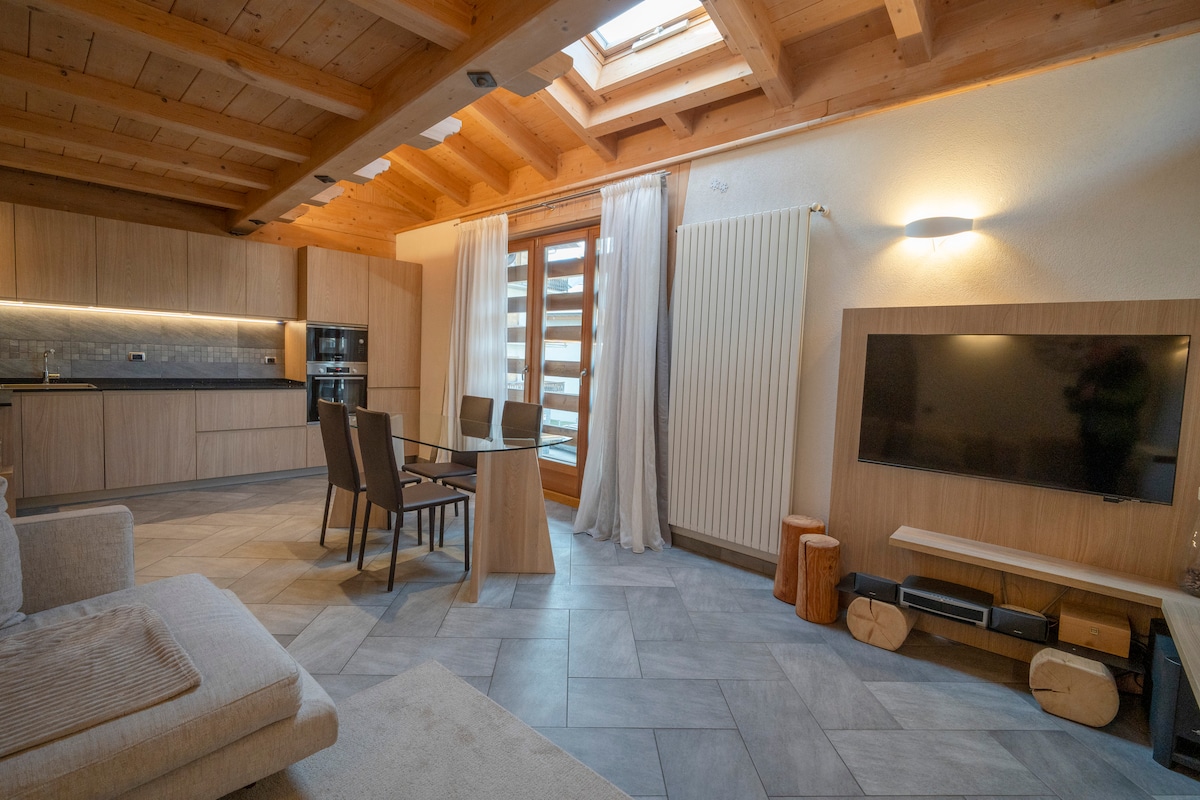 A cozy living area is presented, featuring wooden beams and large windows. The space includes a comfortable sofa, a dining table with chairs, and a modern kitchen with sleek cabinetry. A mounted TV is positioned on the wall opposite the seating area.