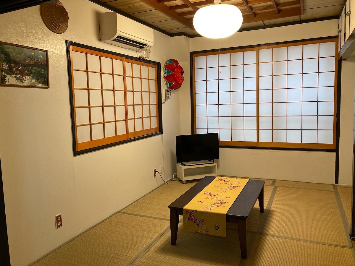 A traditional Japanese-style room is presented, featuring tatami flooring and sliding shoji windows that allow natural light to filter in. A low wooden table is centered on the floor, complemented by a flat-screen television on a nearby stand. Decorative elements include a traditional lantern.