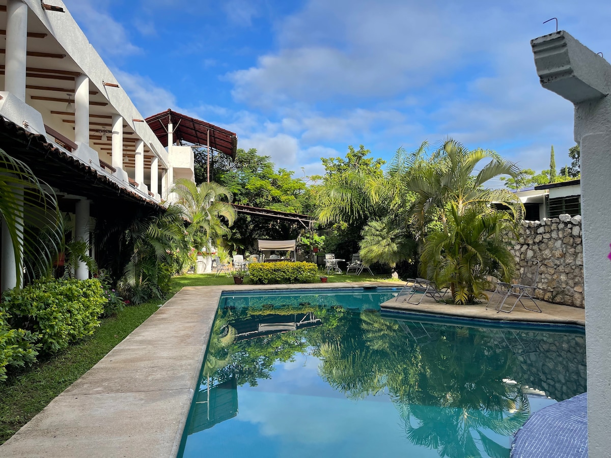 A tranquil swimming pool reflects the surrounding greenery and sky. Lush palm trees and ornamental plants frame the pool area, while lounge chairs are positioned nearby. A covered patio extends along the building, providing shaded spots for relaxation.