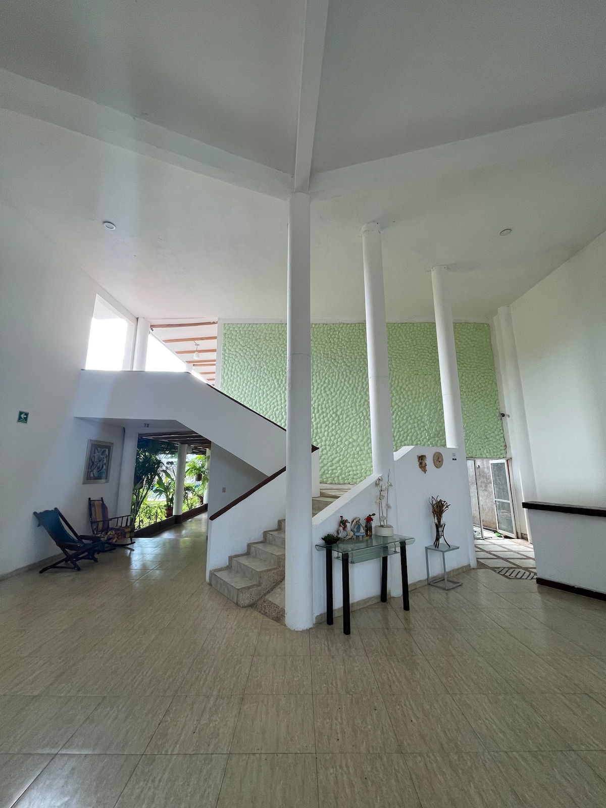 A spacious entrance area is characterized by high ceilings and natural light. An elegant staircase leads to the upper level, while a patterned green wall adds a touch of color. A small table with decorative items is positioned near the base of the stairs.