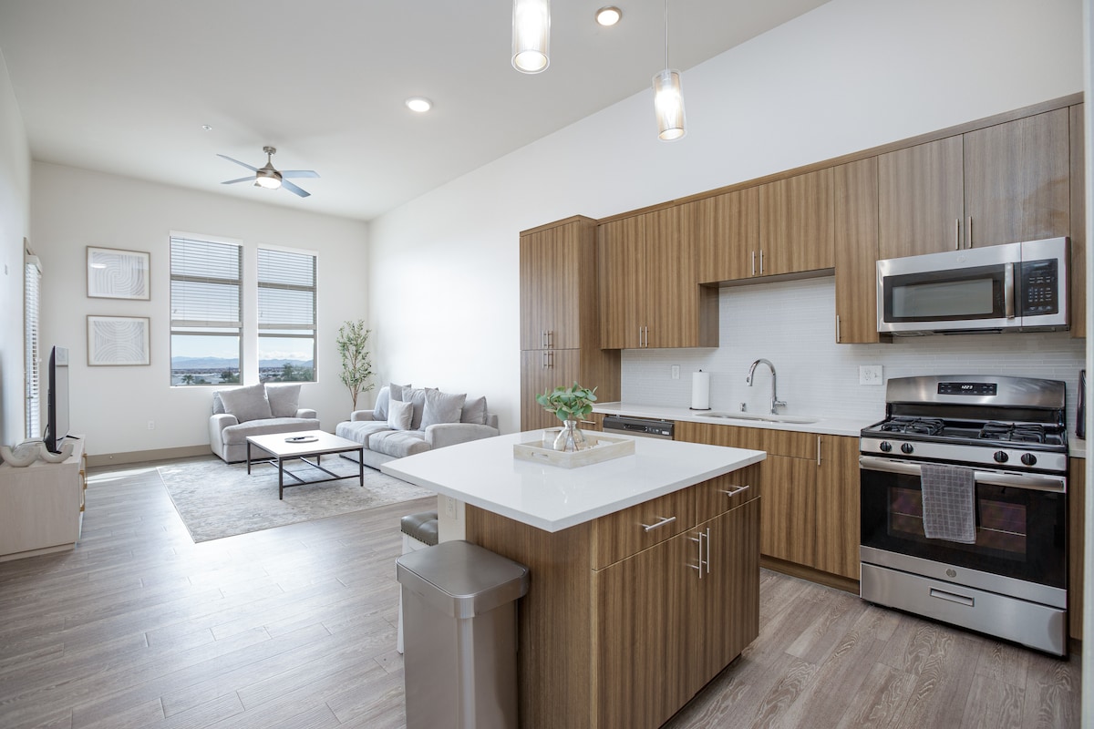 A modern kitchen features sleek wooden cabinets and stainless steel appliances, including a gas stove and microwave. An island with seating invites casual dining, while large windows allow natural light to brighten the open living area, which includes a comfortable seating arrangement.
