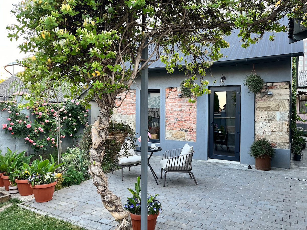 A charming outdoor patio area features stone and brick walls, surrounded by planters filled with greenery and blooming flowers. Two comfortable chairs are positioned near the entrance, which is framed by large glass doors that invite natural light into the space.