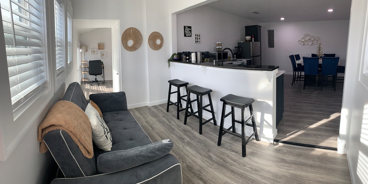 A spacious living area features a comfortable gray couch with decorative pillows. A bar with three stools separates the living space from the kitchen, where dark cabinets and modern appliances are visible. Natural light fills the room through large windows adorned with white shutters.