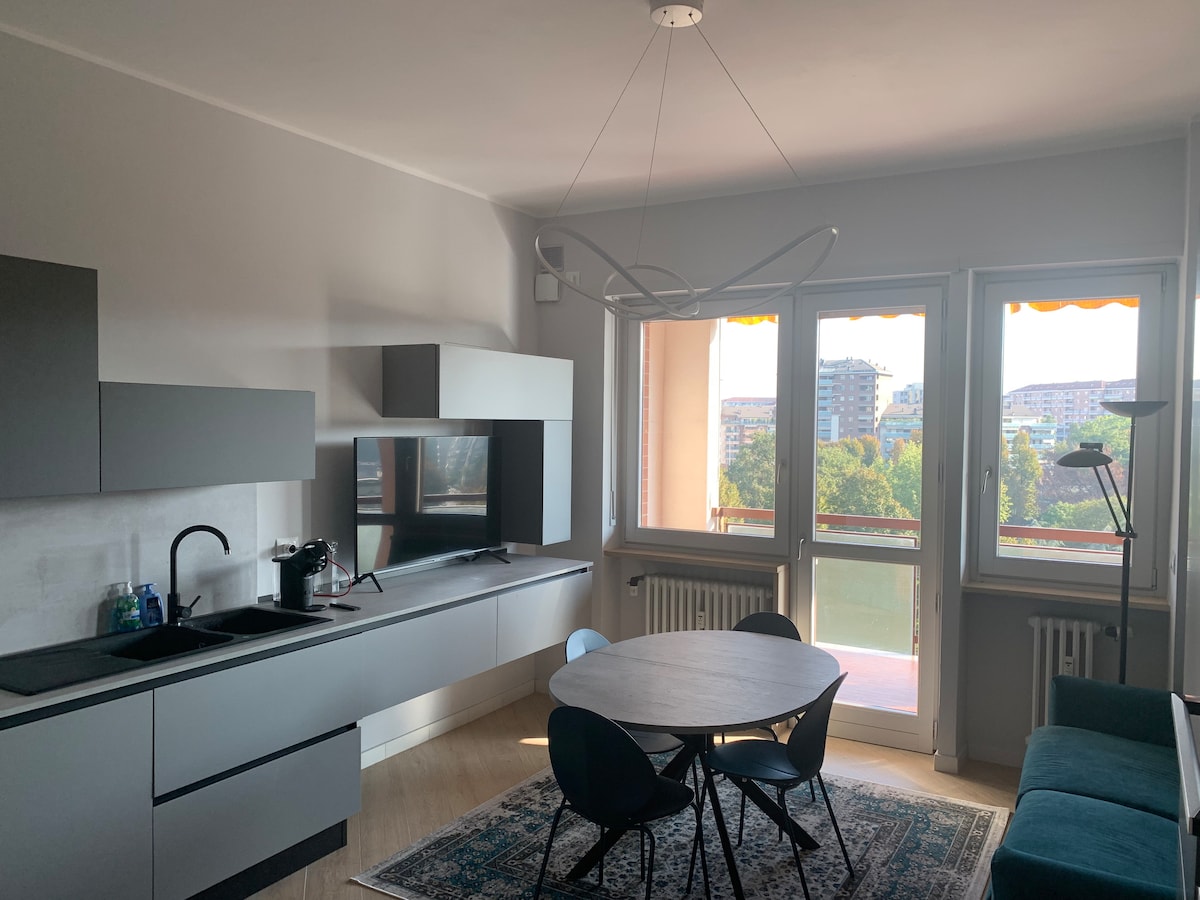 A modern kitchen area features sleek, gray cabinetry and a mounted television. A round dining table with blue chairs is positioned under a decorative light fixture. Large windows allow for ample natural light, offering views of the surrounding landscape.