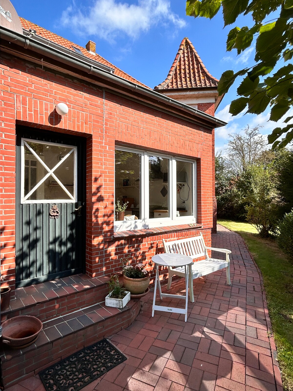 The entrance of a charming brick building features a dark door with a window and a small step leading to a pathway. A white bench is positioned beside the door, surrounded by potted plants. Lush greenery can be seen in the background under a clear blue sky.
