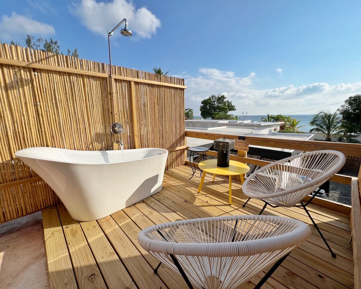 A private rooftop deck features a freestanding tub and two stylish chairs next to a small table. Bamboo fencing encloses the space, providing privacy and a serene atmosphere with ocean views in the background.
