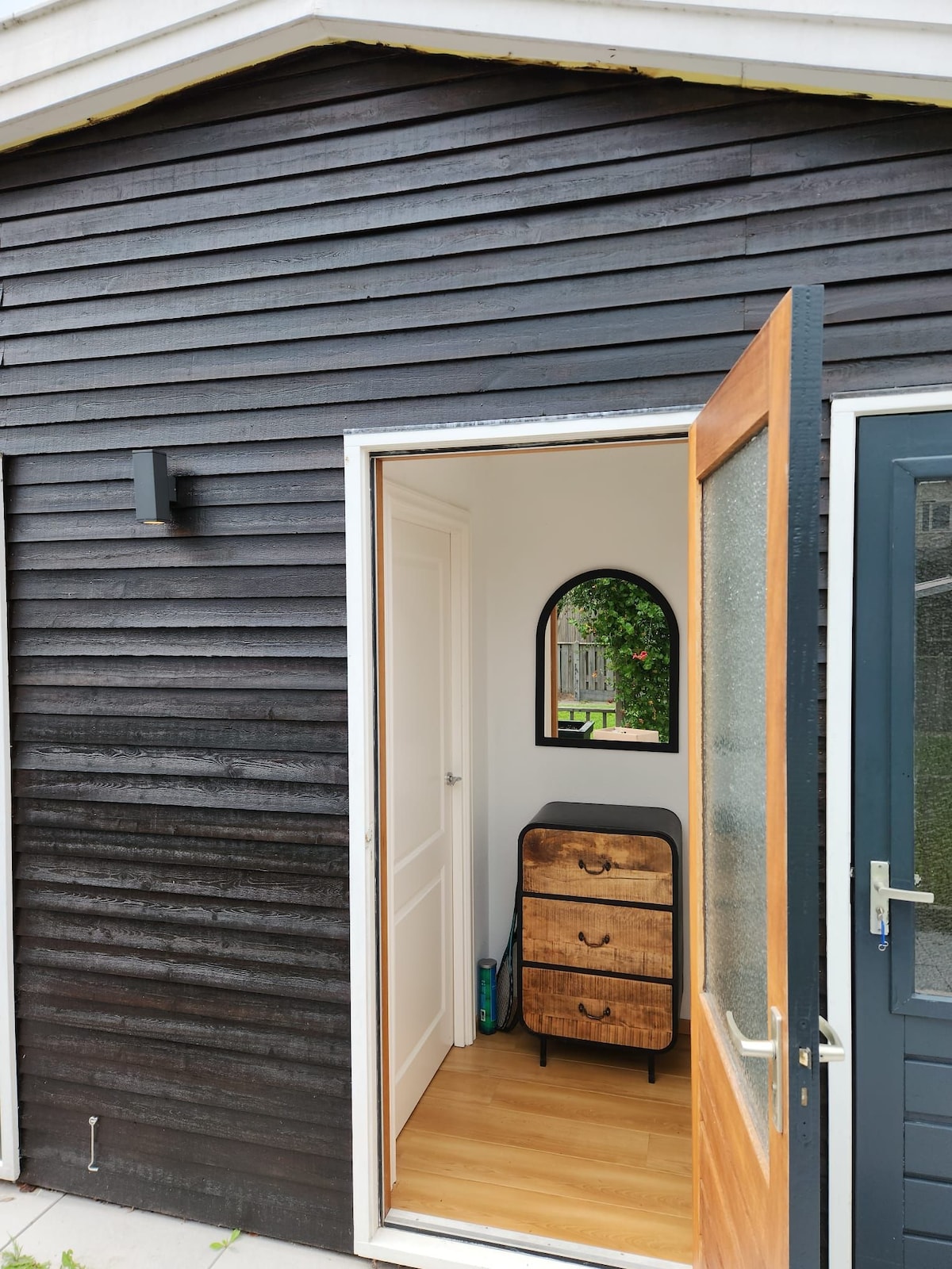 An entrance is visible, featuring a wooden door that is ajar. A small cabinet with a mirror above it can be seen inside, surrounded by walls made of dark wood. Light wooden flooring adds warmth to the space.