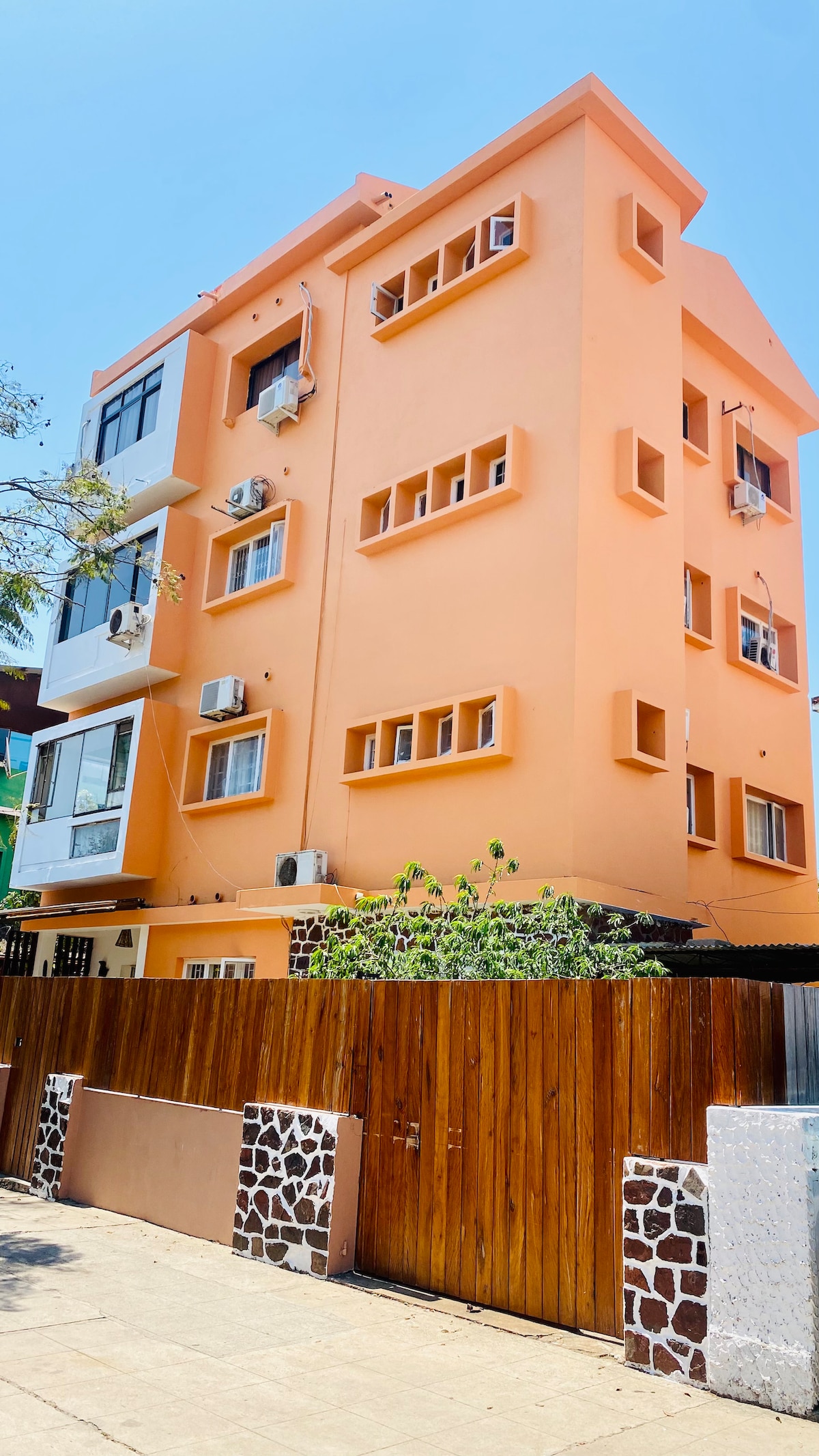 The exterior of a four-story building is presented, featuring orange-painted walls and a number of square windows. Several air conditioning units are mounted on the facade. A wooden gate leads to the entrance, bordered by stone wall accents.