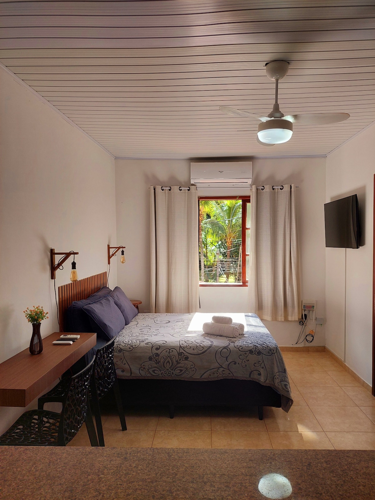 A cozy sleeping area features a double bed with a patterned blanket and soft pillows. Natural light enters through a window adorned with light curtains, revealing greenery outside. A small table and chairs are positioned against the wall, and a television is mounted for convenience.