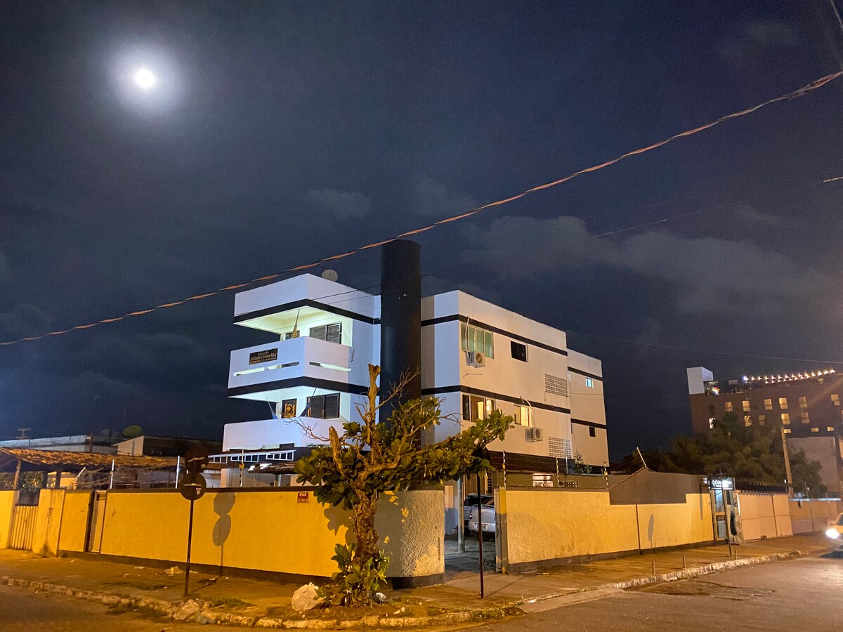 The building is presented from a street view at night, illuminated by the moonlight. Its modern architectural design features multiple levels and balconies. Soft light glows from the windows, highlighting the structured façade against a cloudy sky.