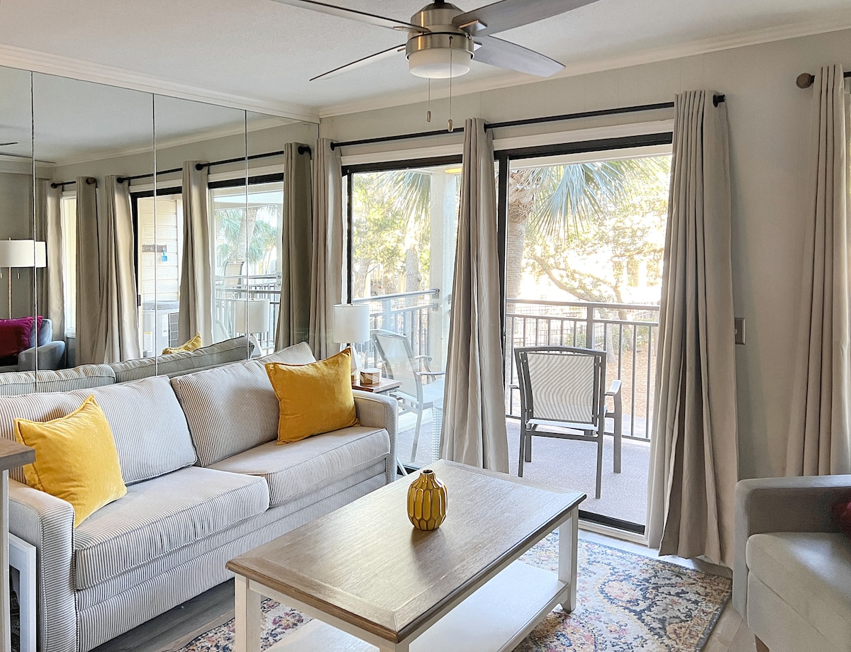 A comfortable living area showcases a light gray sofa adorned with yellow accent pillows. A wooden coffee table sits centered on a patterned rug. Large glass doors allow natural light to fill the space, leading to a balcony surrounded by palm trees.