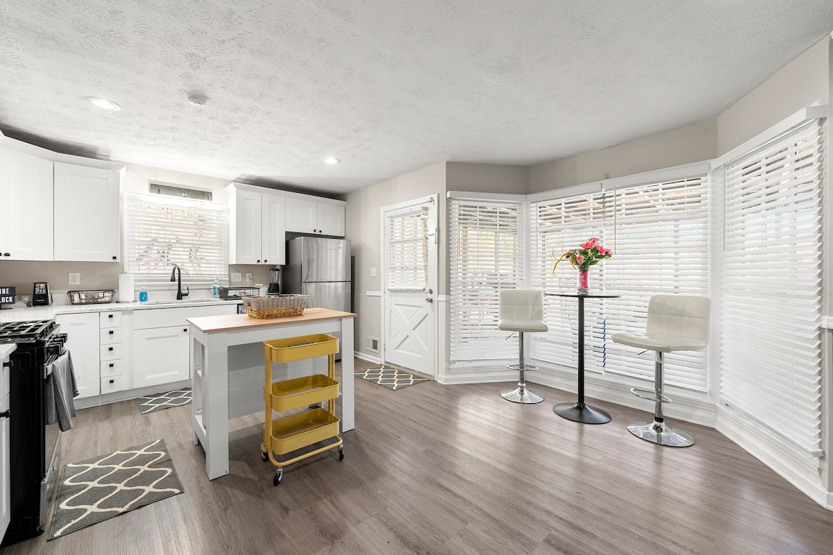 The kitchen area features modern white cabinetry and ample counter space. A central island with seating offers additional prep space. Natural light floods the room through multiple windows adorned with blinds, highlighting a welcoming atmosphere. An entrance door leads to the outdoor area.