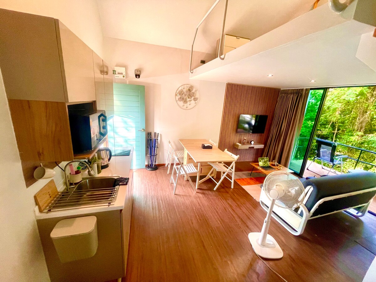 An open living area features a compact kitchen with modern appliances and a dining table surrounded by four chairs. Large windows invite natural light, with a view of greenery. A comfortable sofa is positioned nearby, complemented by a small flat-screen TV on the wall.