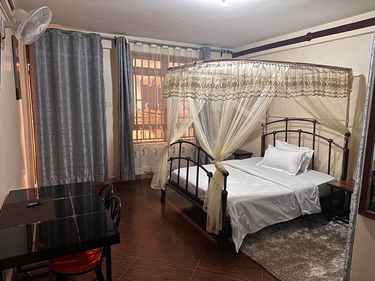 A spacious sleeping area features a canopy bed with sheer curtains, creating a soft, inviting space. Natural light filters through the large window, flanked by elegant drapes. A small dining table is set nearby, offering a convenient workspace.