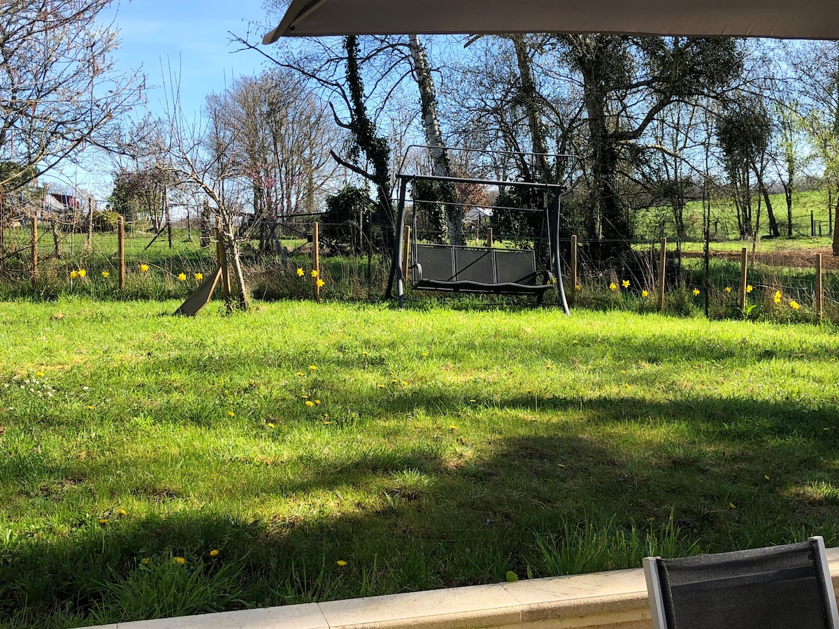 An enclosed garden is visible, featuring a grassy area with a swing seat in the center. Surrounding trees and a fence provide a sense of privacy. Yellow flowers dot the lawn, contributing to the serene rural atmosphere.