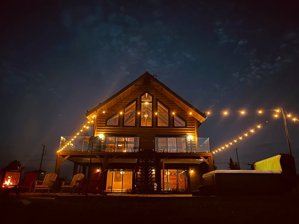 A modern chalet is illuminated at night by soft, warm string lights. The spacious structure features large glass panels that showcase the interior. An outdoor hot tub and fire pit are visible, along with Adirondack chairs arranged near the cozy outdoor fire.