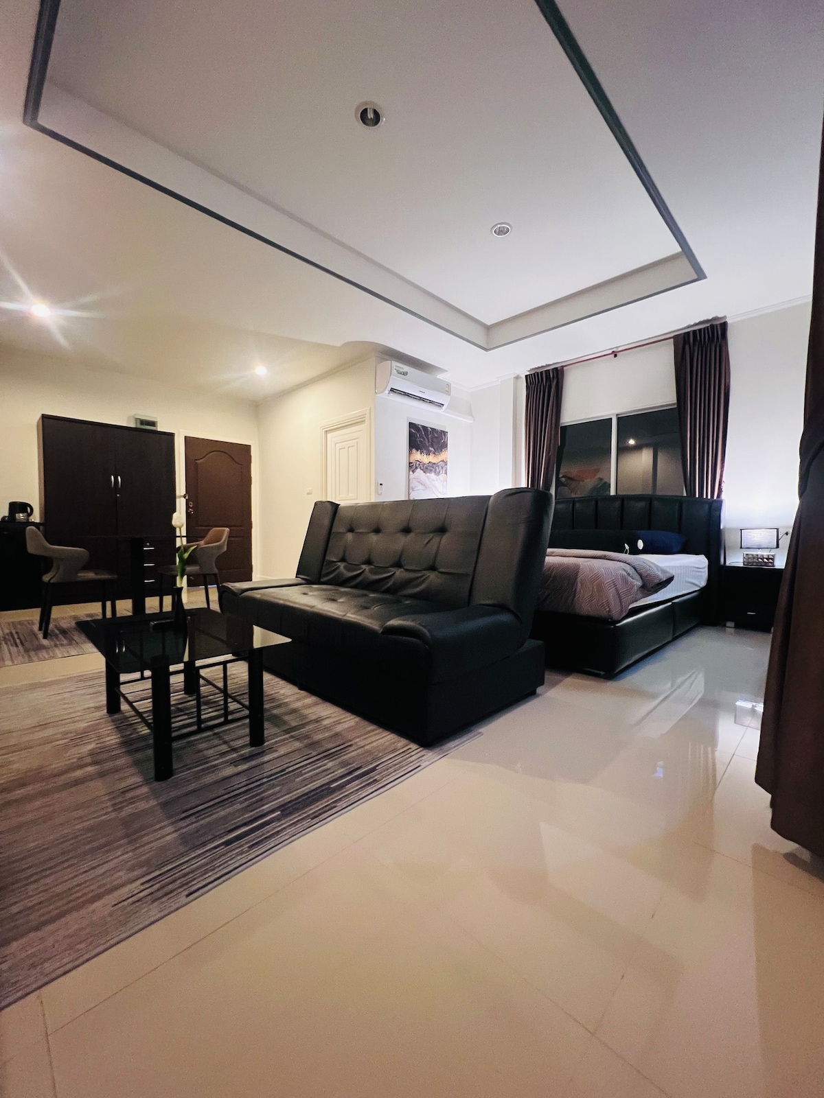 The room features a spacious bed with dark headboard and crisp linens. A modern black sofa sits opposite a compact dining table and chairs. Large windows with dark curtains provide natural light, while the glossy floor adds a touch of elegance.