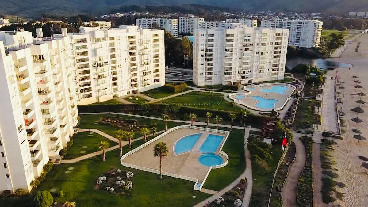 An aerial view showcases multiple buildings within a residential complex, featuring well-maintained landscaped areas. Two swimming pools are visible, with one shaped like a guitar, surrounded by palm trees. The beach and recreational areas are situated nearby, offering easy access to outdoor activities.