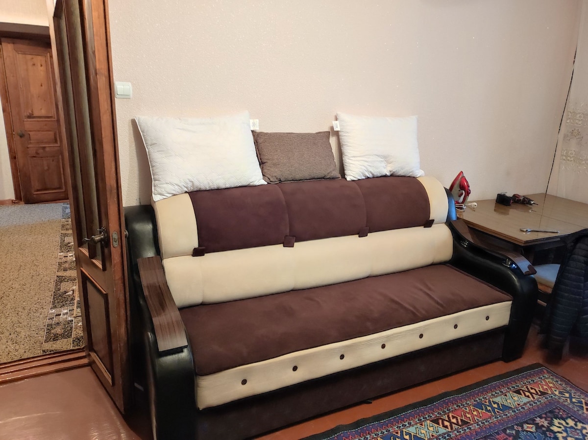 A multifunctional sofa features a brown and cream upholstery, providing a comfortable seating area. Pillows are arranged atop the backrest for added support. A wooden accent is visible on the armrest, and a dining table can be seen in the background, contributing to the cozy layout.